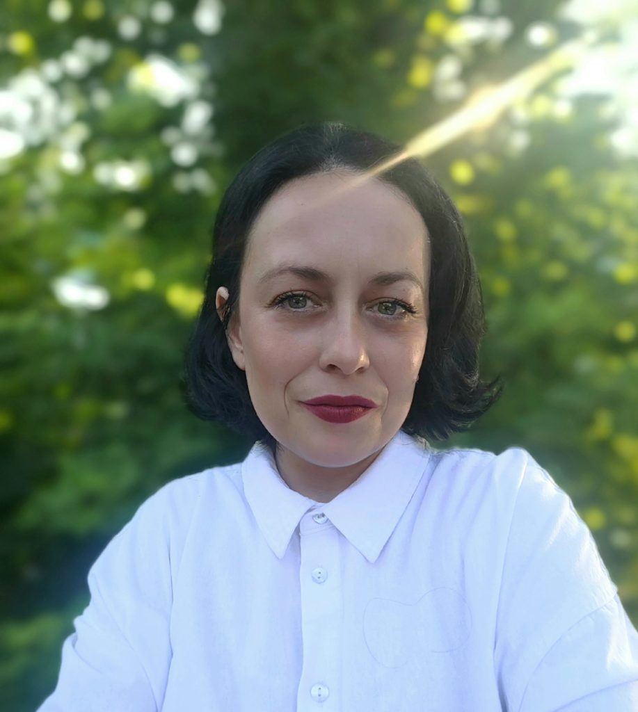 Headshot of Viktória Belle, wearing a white shirt in front of greenery.
